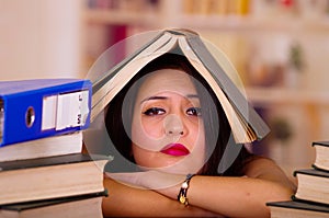 Young brunette woman wearing pink top lying bent over desk with stack of books, open book placed on head, tired facial