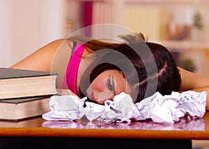 Young brunette woman wearing pink top lying bent over desk with stack of books placed on it, wasted papers around