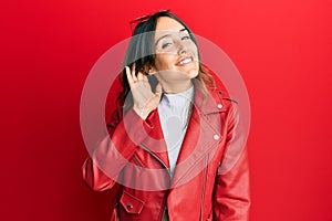 Young brunette woman wearing red leather jacket smiling with hand over ear listening an hearing to rumor or gossip