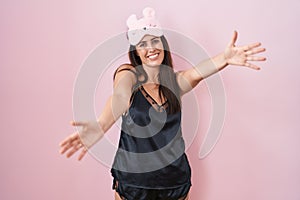 Young brunette woman wearing sleep mask and pyjama looking at the camera smiling with open arms for hug