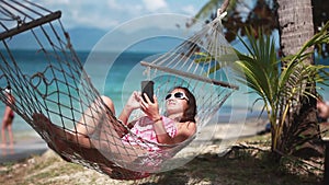 Young brunette woman wearing sunglasses reading sms texting on smartphone lying on hammock relaxing on tropical vacation