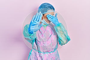 Young brunette woman wearing surgeon uniform and medical mask covering eyes with hands and doing stop gesture with sad and fear