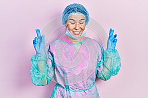 Young brunette woman wearing surgeon uniform and medical mask gesturing finger crossed smiling with hope and eyes closed