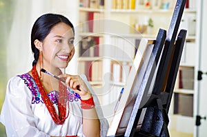 Young brunette woman wearing traditional andean clothing painting on canvas inside studio, evaluating work thoughtful