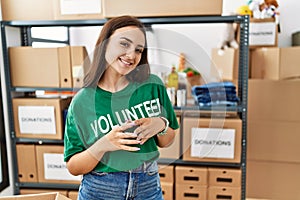 Young brunette woman wearing volunteer t shirt at donations stand hands together and fingers crossed smiling relaxed and cheerful