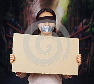 Young brunette woman wearing white sweater, blindfolded black rope and gagged with duct tape, holding up blank sign