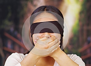 Young brunette woman wearing white sweater, blindfolded with black textile, covering mouth using hands, facing camera