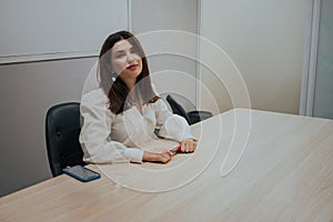 Young brunette woman in a white blouse sitting at the table in the office, holding a marker in her hands and looking at