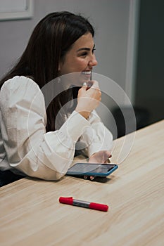 Young brunette woman in a white blouse sitting at the table in the office, holding a smartphone in her hands and smiling