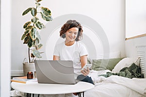 Young brunette woman in white t-shirt using laptop with mobile phone working from home