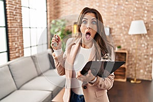Young brunette woman working as real state agent holding keys of new home afraid and shocked with surprise and amazed expression,
