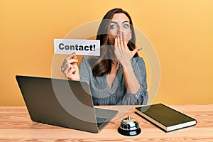 Young brunette woman working at hotel reception holding contact paper covering mouth with hand, shocked and afraid for mistake