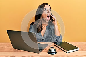 Young brunette woman working at hotel reception talking on the phone angry and mad screaming frustrated and furious, shouting with