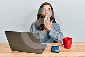Young brunette woman working at the office using computer laptop covering mouth with hand, shocked and afraid for mistake
