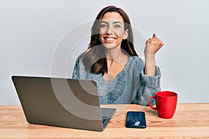 Young brunette woman working at the office using computer laptop screaming proud, celebrating victory and success very excited