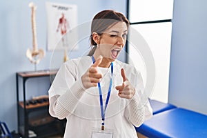 Young brunette woman working at pain recovery clinic pointing fingers to camera with happy and funny face