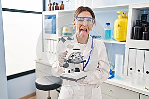 Young brunette woman working at scientist laboratory with microscope sticking tongue out happy with funny expression
