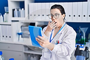 Young brunette woman working at scientist laboratory using tablet covering mouth with hand, shocked and afraid for mistake