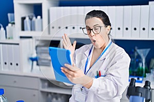 Young brunette woman working at scientist laboratory using tablet scared and amazed with open mouth for surprise, disbelief face