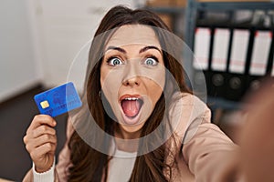 Young brunette woman working at small business ecommerce holding credit card afraid and shocked with surprise and amazed