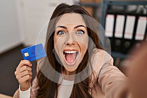 Young brunette woman working at small business ecommerce holding credit card celebrating crazy and amazed for success with open