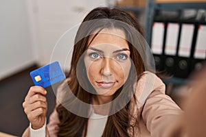 Young brunette woman working at small business ecommerce holding credit card relaxed with serious expression on face