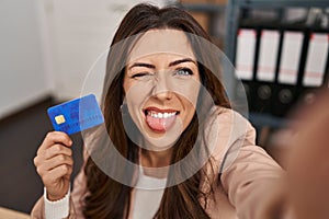 Young brunette woman working at small business ecommerce holding credit card sticking tongue out happy with funny expression