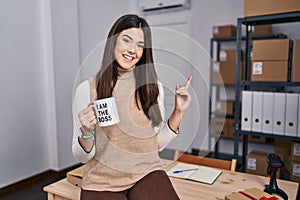 Young brunette woman working at small business ecommerce holding i am the boss cup smiling happy pointing with hand and finger to