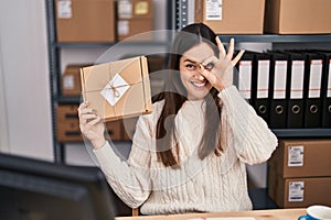 Young brunette woman working at small business ecommerce smiling happy doing ok sign with hand on eye looking through fingers