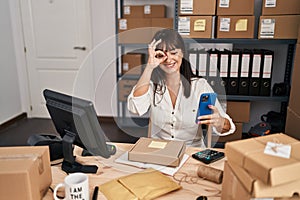 Young brunette woman working at small business ecommerce using smartphone smiling happy doing ok sign with hand on eye looking