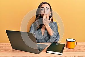 Young brunette woman working using computer laptop covering mouth with hand, shocked and afraid for mistake