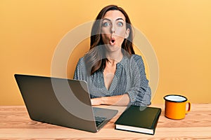 Young brunette woman working using computer laptop scared and amazed with open mouth for surprise, disbelief face