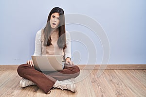 Young brunette woman working using computer laptop sitting on the floor in shock face, looking skeptical and sarcastic, surprised