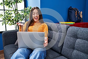 Young brunette woman working using computer laptop sitting on the sofa celebrating achievement with happy smile and winner