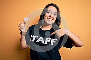 Young brunette worker woman wearing staff t-shirt as uniform showing puzzle piece as teamwork with surprise face pointing finger