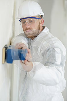 Young builder in hardhat sanding wall indoors