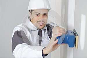 Young builder in hardhat sanding wall indoors