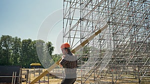 Young builder in helmet carrying heavy metal plank, working near huge installation on straw field