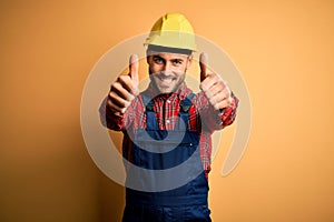 Young builder man wearing construction uniform and safety helmet over yellow isolated background approving doing positive gesture