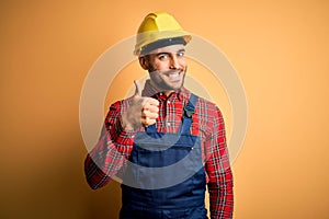 Young builder man wearing construction uniform and safety helmet over yellow isolated background doing happy thumbs up gesture