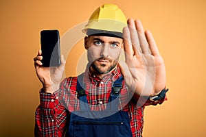 Young builder man wearing safety helmet showing smartphone screen over yellow background with open hand doing stop sign with