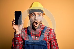 Young builder man wearing safety helmet showing smartphone screen over yellow background scared in shock with a surprise face,