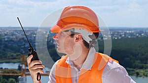 Young builder man wearing safety helmet and using walkie-talkie, discussing, standing outdoors. Business, building