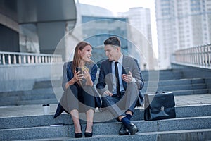 Young business couple with smartphones sitting near office building.