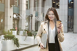 Young business lady holding laptop under arm walking down the street near office center with cup of coffee looking busy, eating on