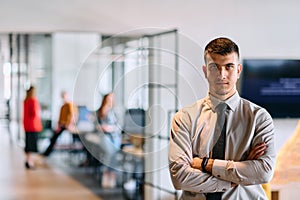 A young business leader stands with crossed arms in a modern office hallway, radiating confidence and a sense of purpose