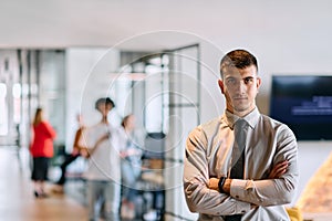 A young business leader stands with crossed arms in a modern office hallway, radiating confidence and a sense of purpose