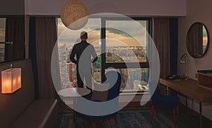 Young business man on the background of the panoramic windows with the view on the city from the hotel room at sunset time