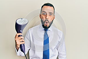 Young business man with beard holding electric steam iron scared and amazed with open mouth for surprise, disbelief face