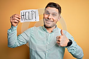 Young business man with blue eyes asking for help holding paper note over yellow background happy with big smile doing ok sign,